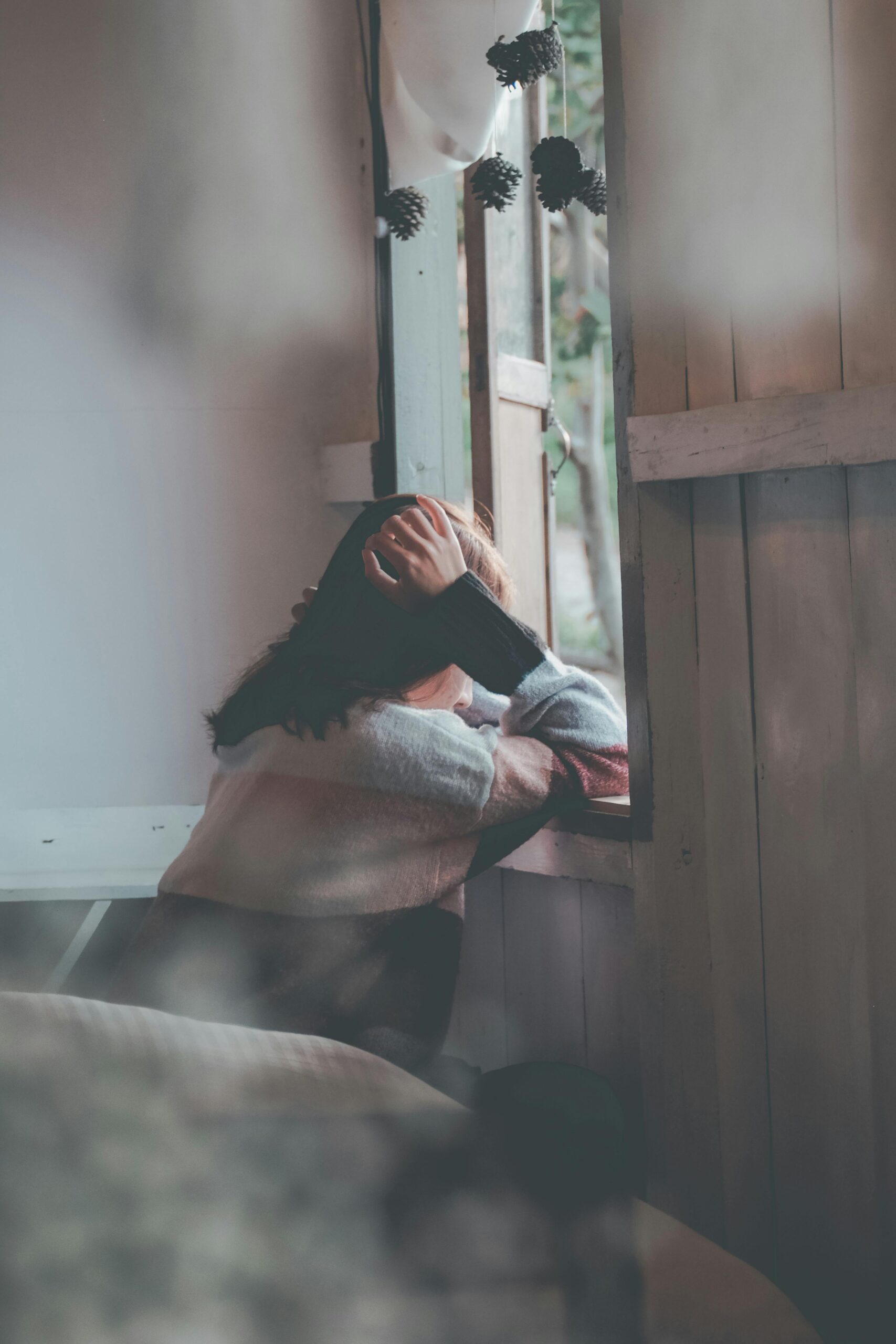 A woman sits indoors gazing thoughtfully out of a window, evoking emotions of loneliness and reflection.