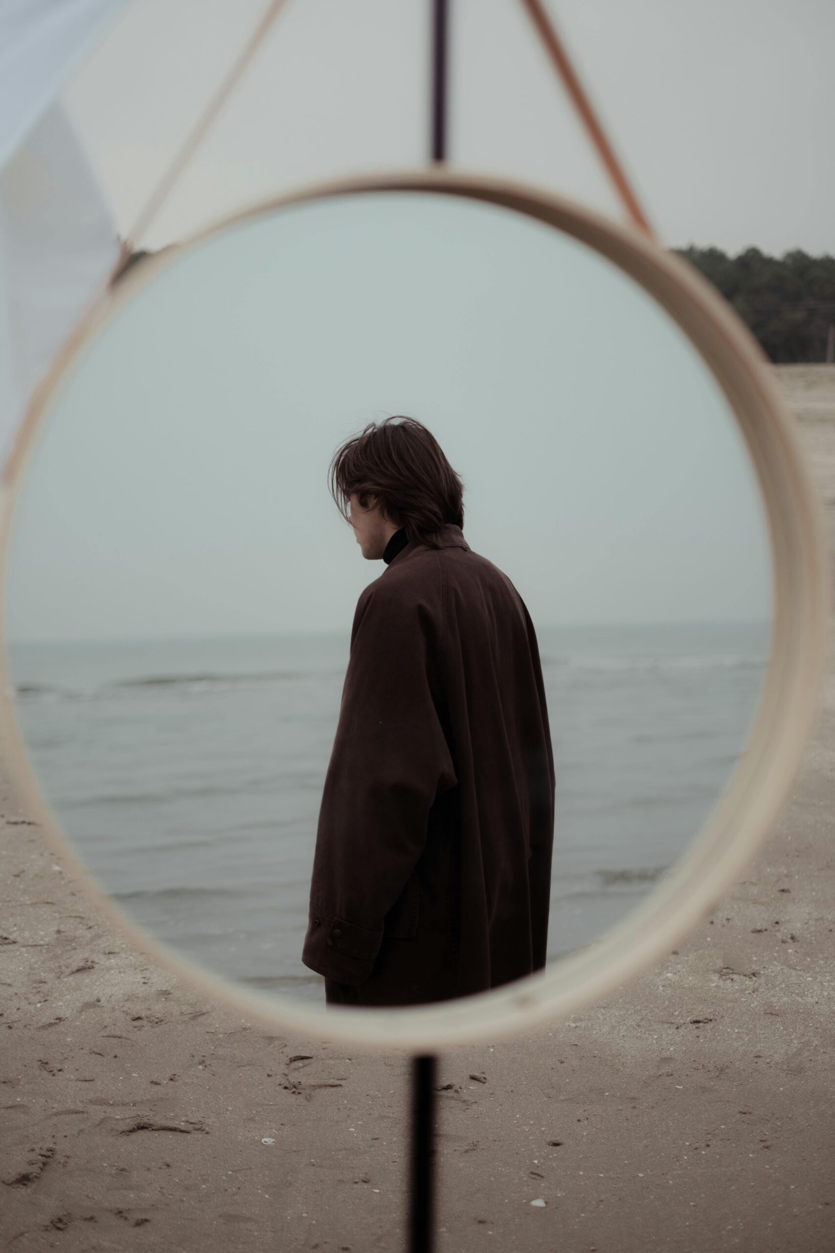 A man in a coat stands alone on the beach, reflected in a round mirror. Moody coastal scene.